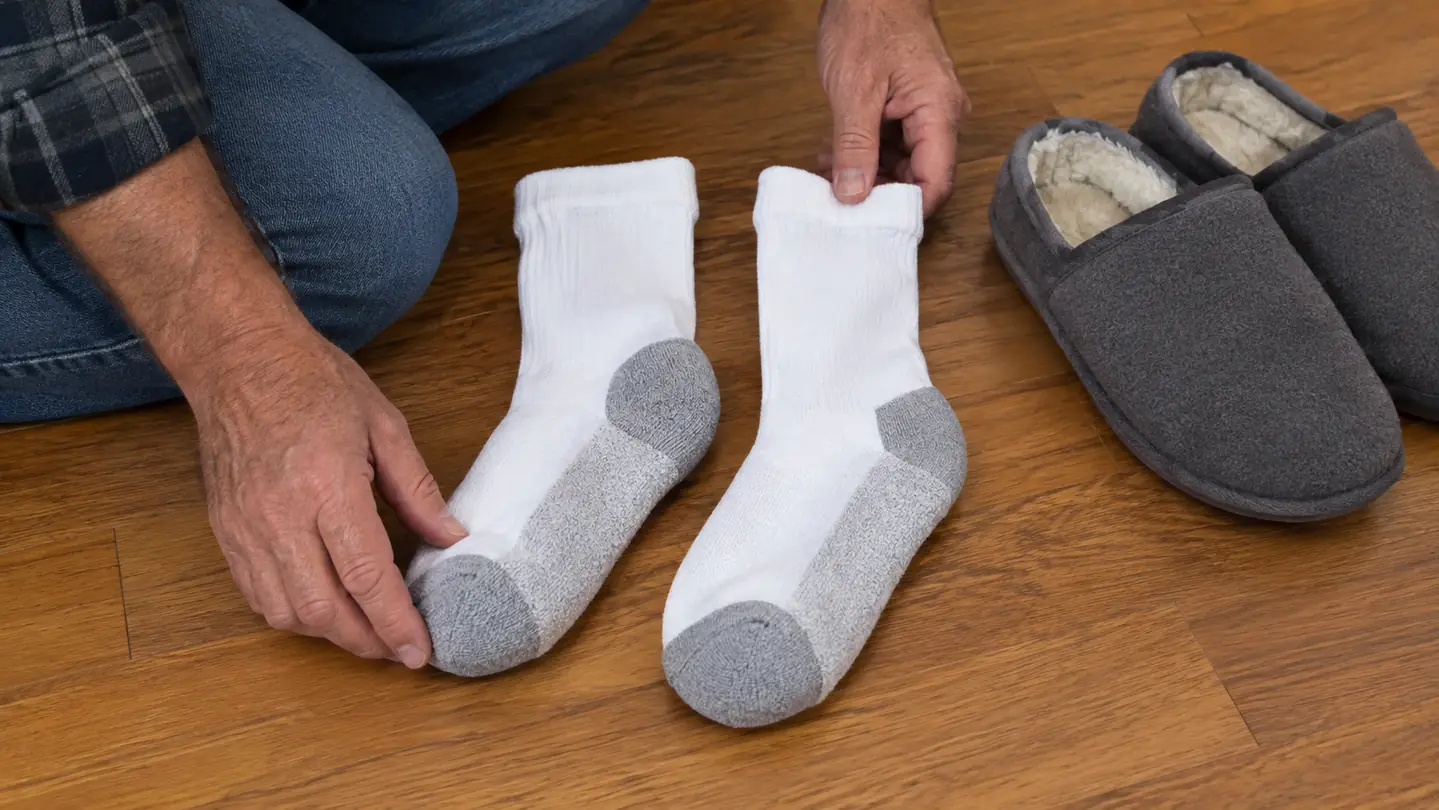 A pair of cushioned non-binding socks laid flat on a hardwood floor beside a pair of slippers